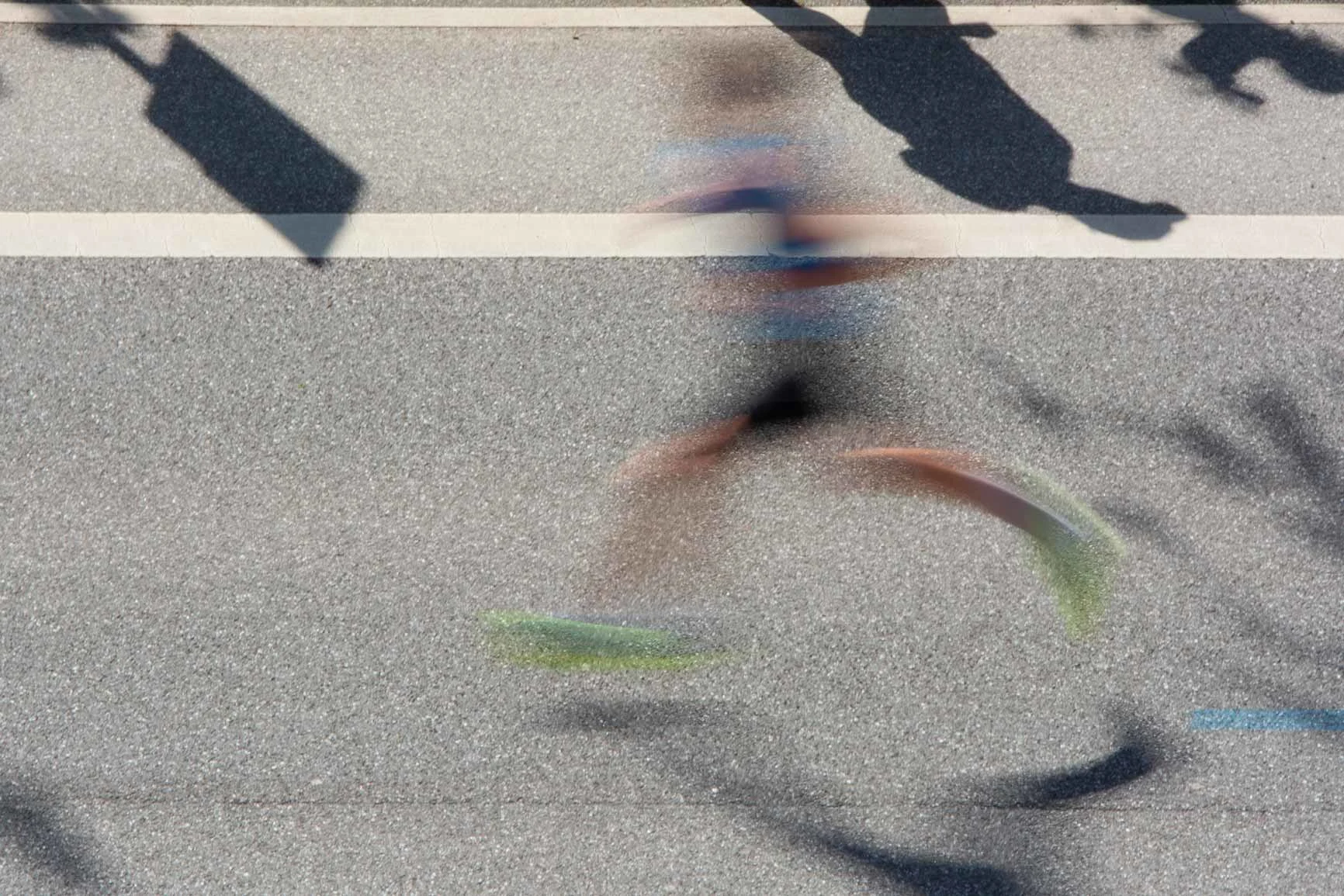 Fotografía borrosa de un corredor con ropa deportiva en una calle con marcas blancas y sombras de transeúntes. Fotografía borrosa de un corredor con ropa deportiva en una calle con marcas blancas y sombras de transeúntes.