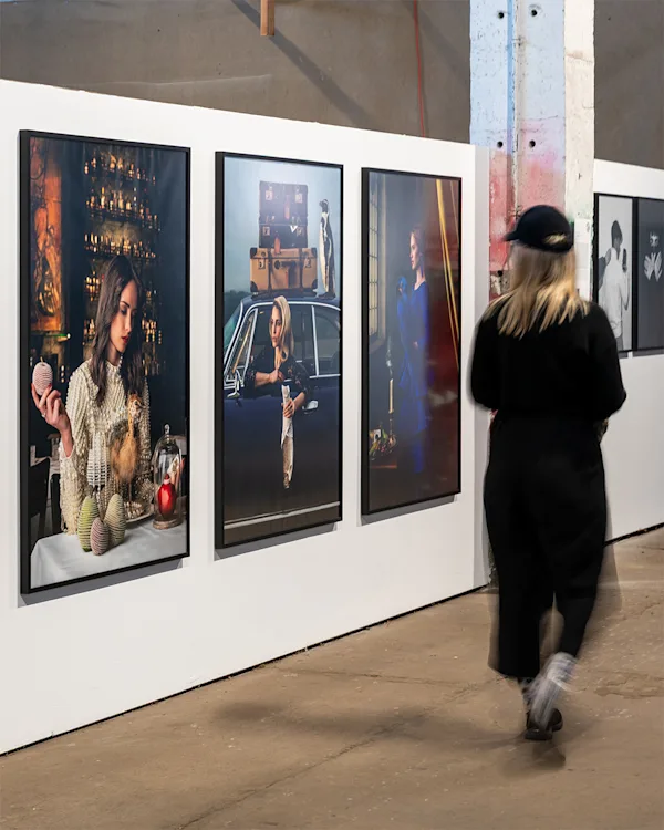 WhiteWall x BFF Scholarship Exhibition A woman in black walks past large framed editorial-style photographs displayed on a white wall in an industrial gallery space.