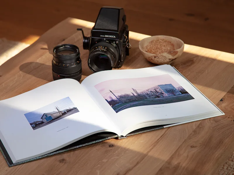 Coffee Table Book An open photo book, a camera, extra lens, and bowl on a wooden table in sunlight.