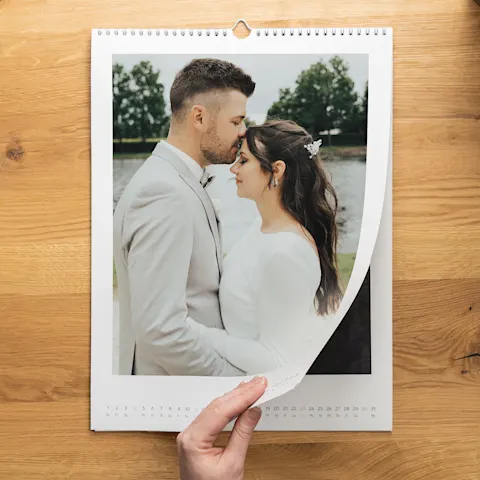 Calendario WhiteWall con foto di matrimonio Una mano che gira la pagina di un calendario da parete a spirale raffigurante una coppia di sposi in abiti formali, uno di fronte all’altra, all’aperto.