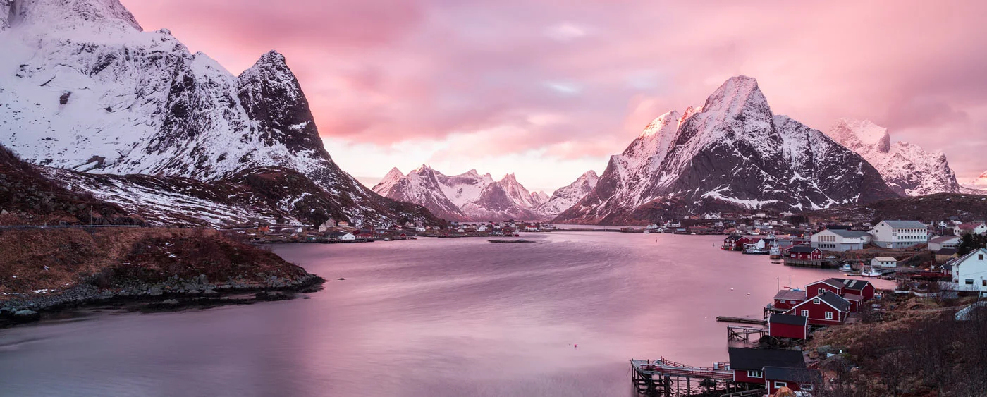 Snow-covered mountains surround a tranquil fjord at sunset with a pink sky and houses on the shore. Snow-covered mountains surround a tranquil fjord at sunset with a pink sky and houses on the shore.