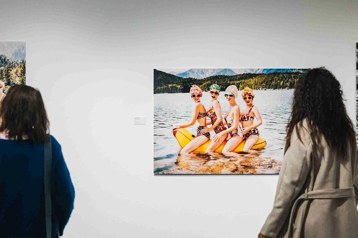 Femmes sur un bateau banane Trois femmes en maillot de bain sont assises sur un bateau banane gonflable jaune dans l'eau, avec des montagnes et un lac en arrière-plan.