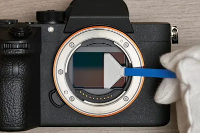 Entretenir son appareil photo 1 Un appareil photo sans objectif montre le capteur. Une main vêtue d'un gant blanc tient une spatule de nettoyage bleue sur le capteur de la caméra.