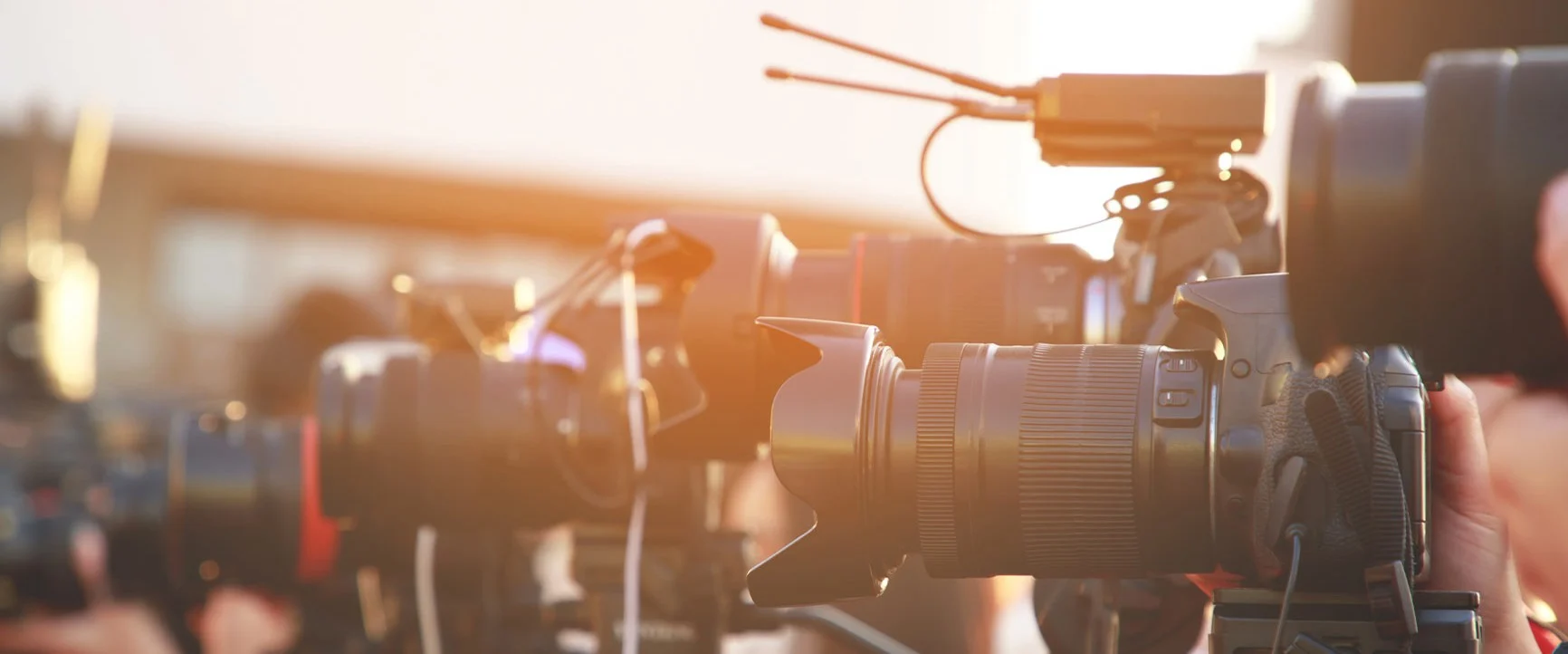 A row of professional cameras mounted on tripods in sunlight, with microphones attached to the cameras.