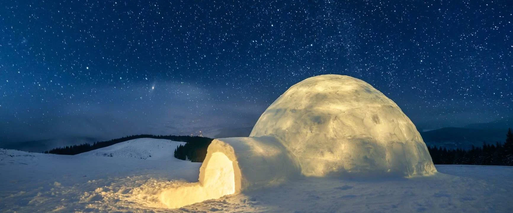 An illuminated snow igloo under a clear starry sky in a snow-covered landscape.
