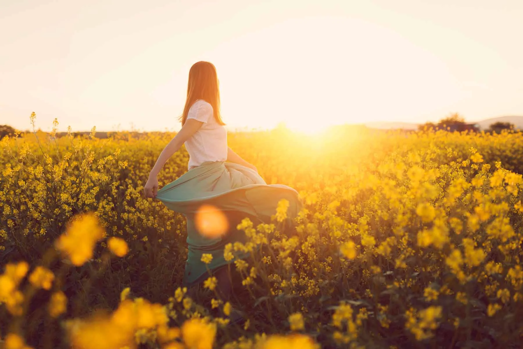 Una mujer con falda está de pie en un campo de colza en flor, con el sol brillando en el cielo al fondo. Una mujer con falda está de pie en un campo de colza en flor, con el sol brillando en el cielo al fondo.