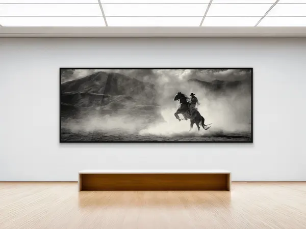 WhiteWall Masterprint Schwarz-weißes gerahmtes Foto eines Cowboys zu Pferd, der durch staubiges Gelände reitet, aufgehängt an einer minimalistischen weißen Galeriewand