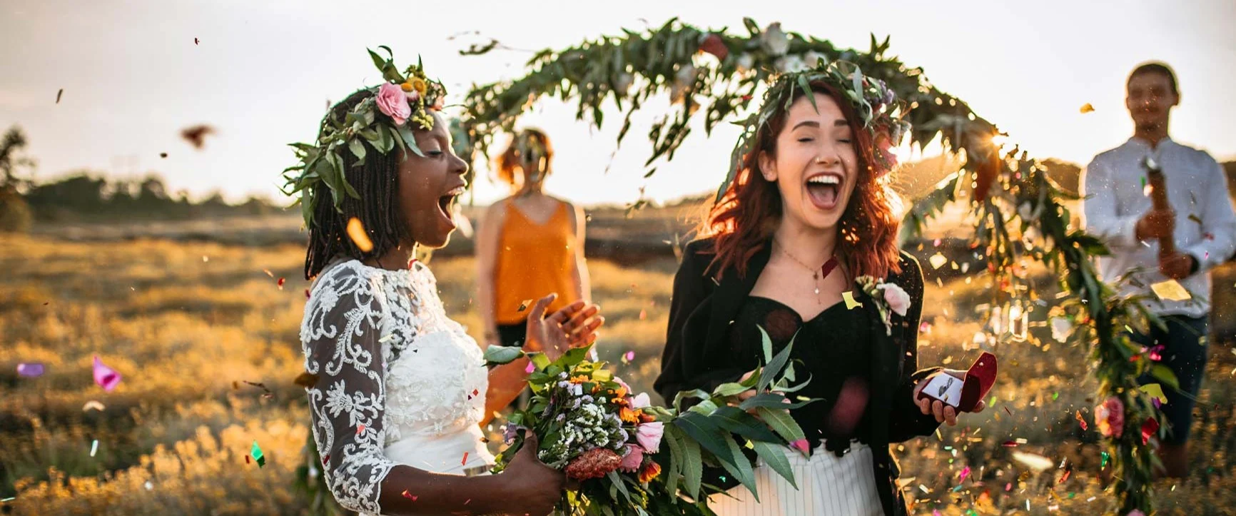 A bride in a white lace dress holding a bouquet, and a woman in a black jacket and white pants, standing in front of a flower-decorated arch outdoors at sunset, with confetti in the background.