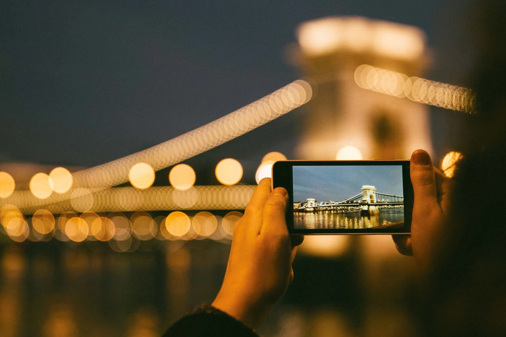 Person photographing the illuminated chain bridge in Budapest at night with a smartphone. Person photographing the illuminated chain bridge in Budapest at night with a smartphone.
