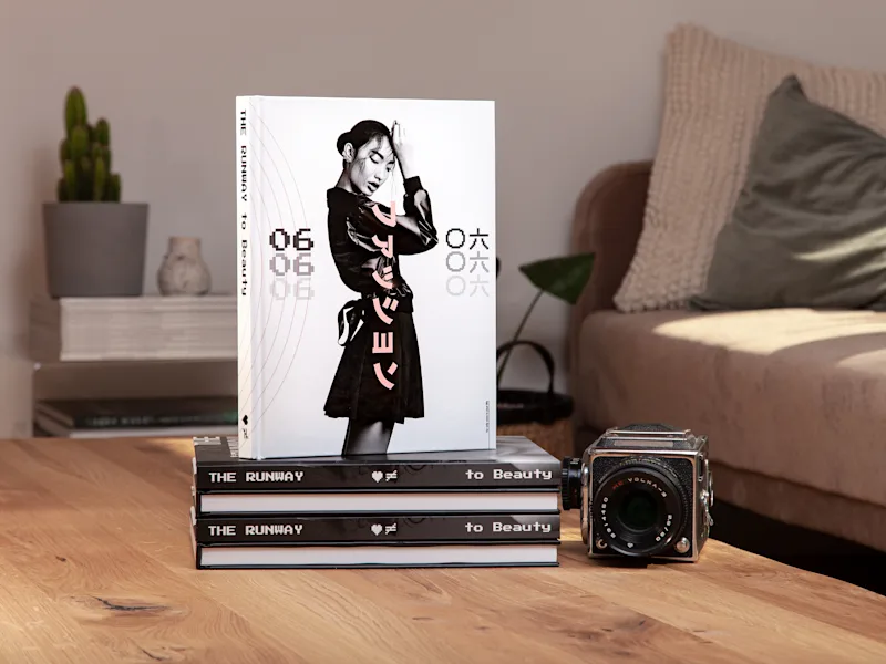 Coffee Table Book A photo book with a model on the cover stands on stacked books near a vintage camera in a cozy living room.