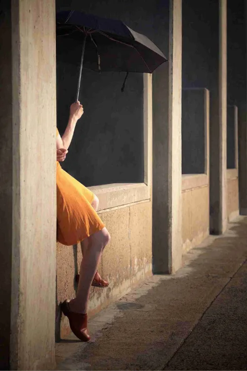 Fine art photography Mania de Praeter An atmospheric fine art photograph by Mania de Praeter depicts a partial view of a person wearing a vibrant orange dress and brown leather clogs. The subject is leaning against a textured concrete wall, partially hidden behind a pillar while holding up a large open black umbrella. A row of brutalist concrete pillars extends into the background, creating a rhythmic sequence of light and shadow. The moody, cinematic lighting accentuates the textures of the stone and the rich color of the fabric.