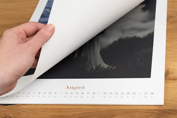 Startmonat des Fotokalenders Hand dreht Seite eines Wandkalenders, um den Monat August mit Daten und Tagen auf hölzernem Hintergrund zu zeigen