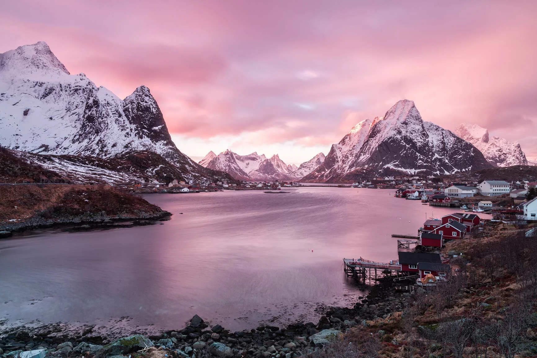 Schneebedeckte Berge und ein ruhiger Fjord bei Sonnenuntergang mit rosa Himmel, kleine Häuser am Ufer. Schneebedeckte Berge und ein ruhiger Fjord bei Sonnenuntergang mit rosa Himmel, kleine Häuser am Ufer.