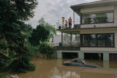 DOCKS Collective - Documentary photography Flooded house with balcony, people in safety vests, partially submerged car in brown water.