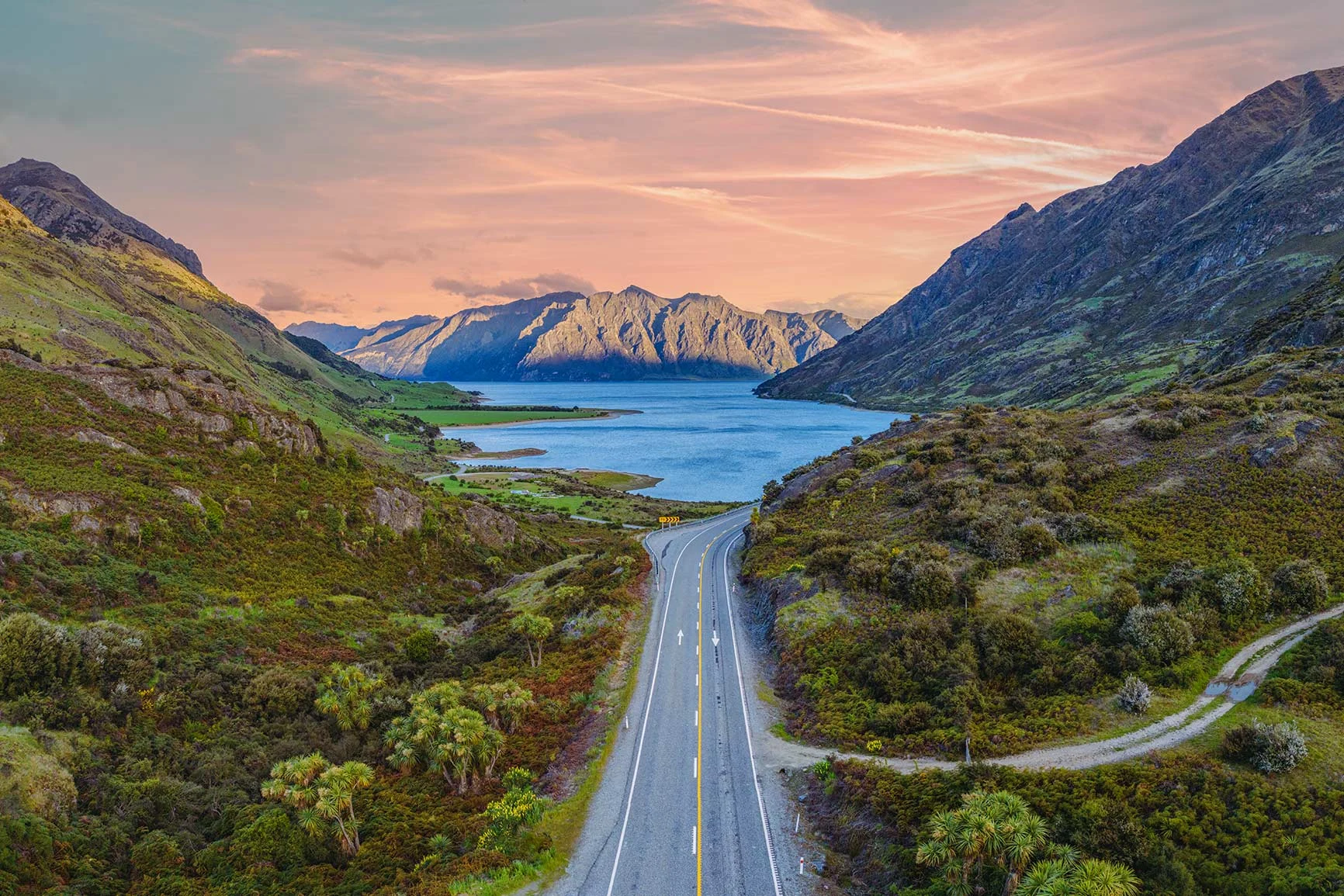 Una strada attraversa verdi colline e conduce a un lago circondato da montagne al tramonto in Nuova Zelanda. Una strada attraversa verdi colline e conduce a un lago circondato da montagne al tramonto in Nuova Zelanda.