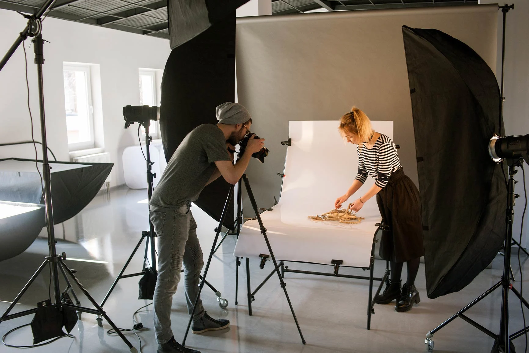 Fotografia di prodotti in studio Studio fotografico con un fotografo che osserva una macchina fotografica montata su un treppiede e una donna che dispone degli oggetti su uno sfondo bianco.