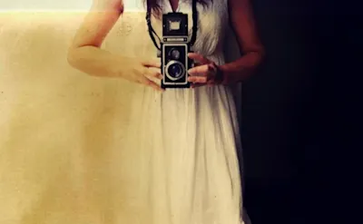 ZEIT Akademie - „Fotografie – Die Kunst der guten Bilder“ Frau in weißem Kleid hält eine Vintage-Kamera vor einem Spiegel.