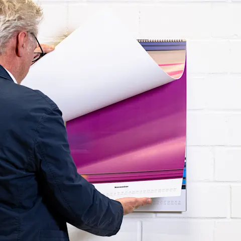 Fotografo con un calendario Un uomo che sfoglia un grande calendario da parete aperto su novembre, con vivaci immagini astratte viola e rosa.