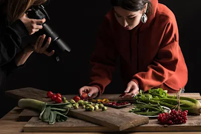 Food photography of vegetables A person in a red sweater is arranging vegetables on a wooden board in front of another person holding a camera in a dark room.