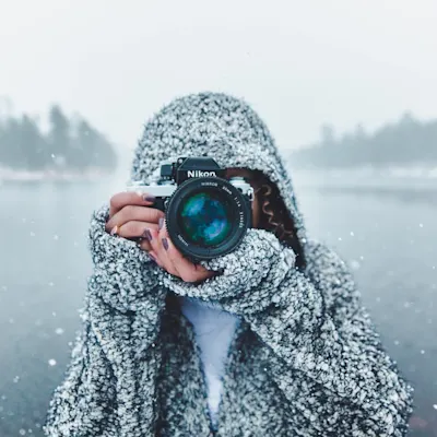 Photographer with a camera as symbol for the magazine Photographer in speckled gray winter coat holds Nikon camera during snowfall, with blue lens reflection and misty background.