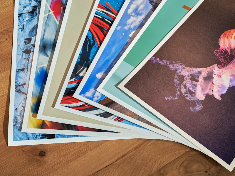 Fine Art Print 2 Various fine art papers with photo prints including jellyfish, cloud sky, and abstract patterns, fanned out on wooden table.