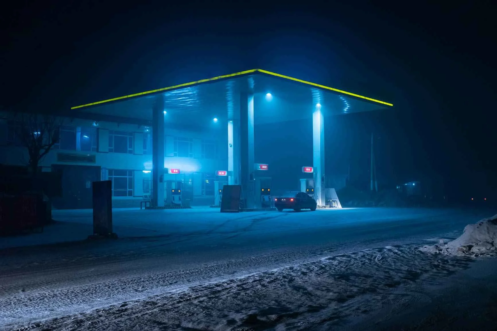 Gas station at night A gas station lit up at night, with three pumps and a snow-covered ground, set against a foggy backdrop.