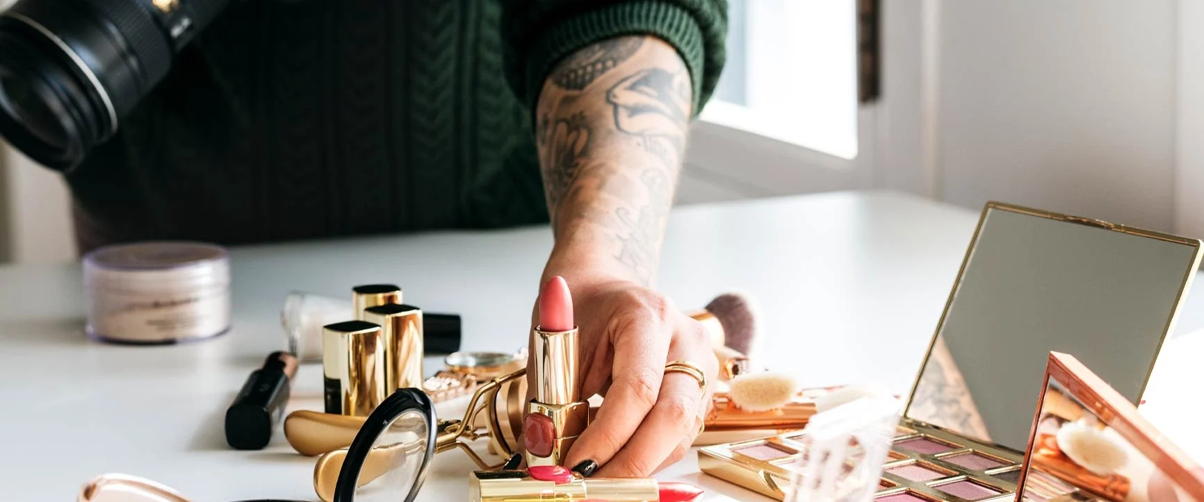 Diversi prodotti di bellezza su un tavolo. Sullo sfondo, una donna con una macchina fotografica sta prendendo un rossetto rosso.