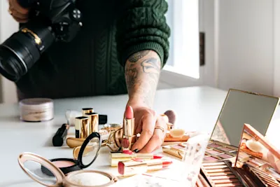 Product photography of cosmetics Various cosmetics on a table. In the background, a woman holding a camera reaches for a red lipstick.