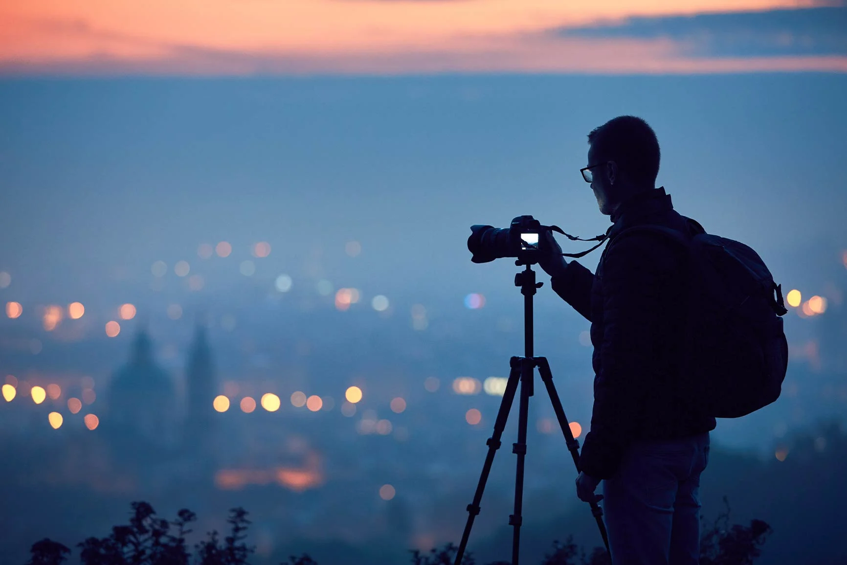 Silhouette of a photographer The silhouette of a photographer with a backpack, operating a camera on a tripod at dusk against a blurred cityscape.