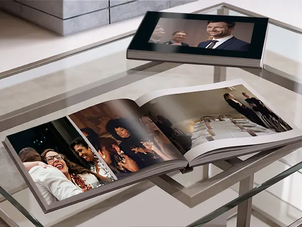 Livre photo haut de gamme Deux livres photo professionnels sur une table en verre : l’un est ouvert et présente des photos d’une célébration de mariage, l’autre est fermé avec le portrait d’un homme sur la couverture.