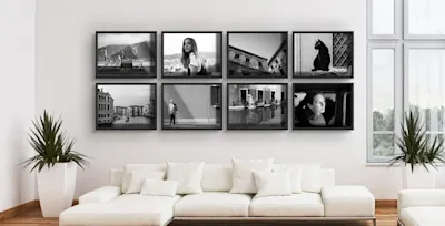 Bilder gekonnt aufhängen 1 Acht schwarz-weiße Bilder in schwarzen Rahmen an Wand über Sofa mit Kissen, zwei Pflanzen seitlich.
