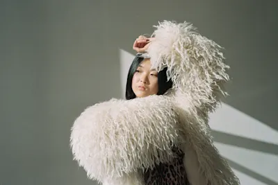 Woman in a fur cloak A woman with black hair and a white, long-haired fur cloak raises one arm against a light-colored background.