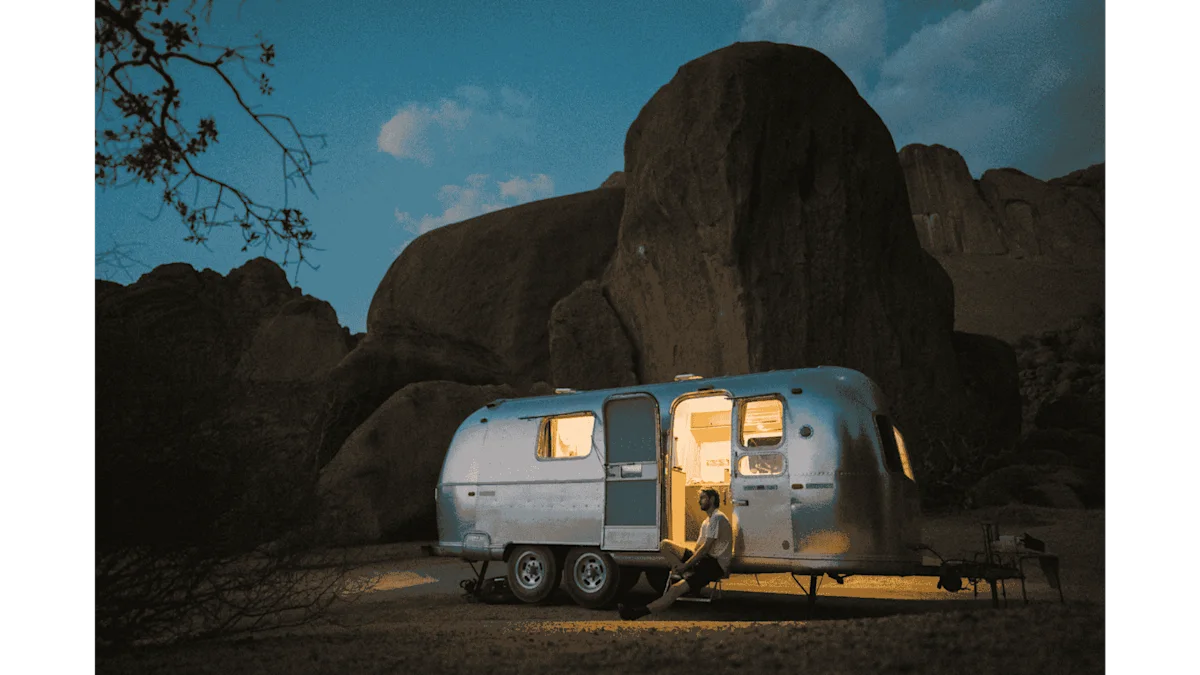 Thylacine en Namibie – Ambiance de soirée au studio mobile Un cliché nocturne plein d'ambiance dans le désert namibien. Une caravane Airstream argentée est vivement éclairée de l'intérieur, projetant une lueur jaune chaleureuse par la porte ouverte et les fenêtres sur le sol sablonneux. Un homme est assis, détendu, dans l'encadrement de la porte du véhicule. À l'arrière-plan, des formations rocheuses sombres et massives s'élèvent sous un ciel nocturne d'un bleu profond et légèrement nuageux.