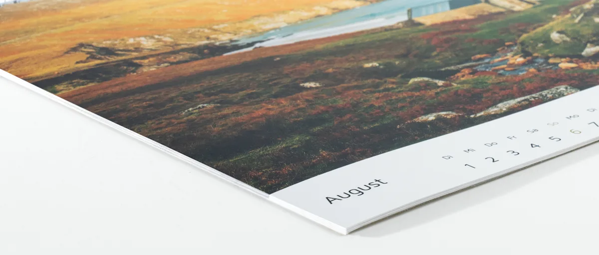 Fotokalender Fotopapier tiefmatt Kalenderseiten aufgefächert, die den August zeigen, mit einer Landschaftsaufnahme in erdigen Braun- und Grüntönen im oberen Bereich