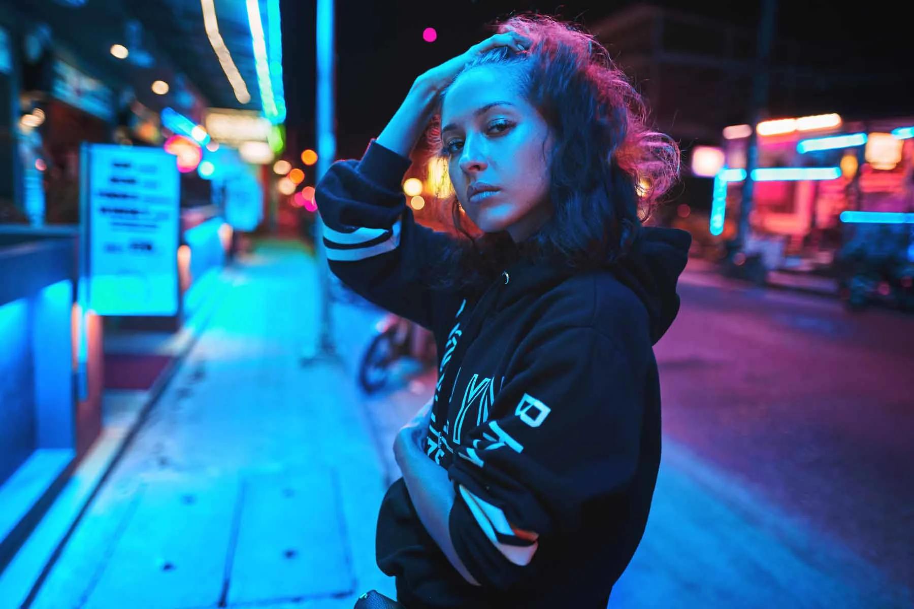 Una persona con sudadera con capucha está de pie por la noche en una acera con iluminación de neón azul y rojo. Una persona con sudadera con capucha está de pie por la noche en una acera con iluminación de neón azul y rojo.