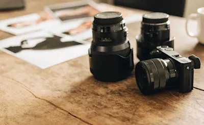 Types de caméras 1 Un appareil photo avec objectif interchangeable et deux objectifs supplémentaires sont posés sur une table en bois. Des tirages photo sont visibles en arrière-plan.