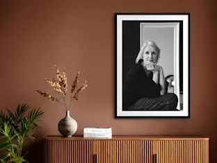 Black and white portrait in a matted frameabove wooden credenza with vase of dried flowers, books.