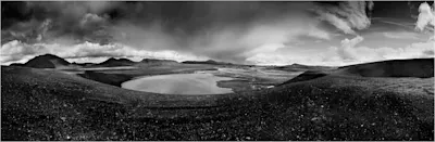 Nina Papiorek – Schwarz-Weiß-Landschaft Weite Landschaft mit See, Hügeln und dramatischer Wolkenformation in Schwarz-Weiß.