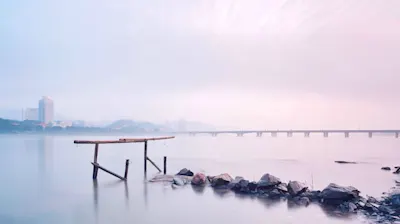 Lignes et perspectives 1 Passerelle en bois et rochers dans l'eau, avec un pont et des bâtiments en arrière-plan, dans la douce lumière du matin.