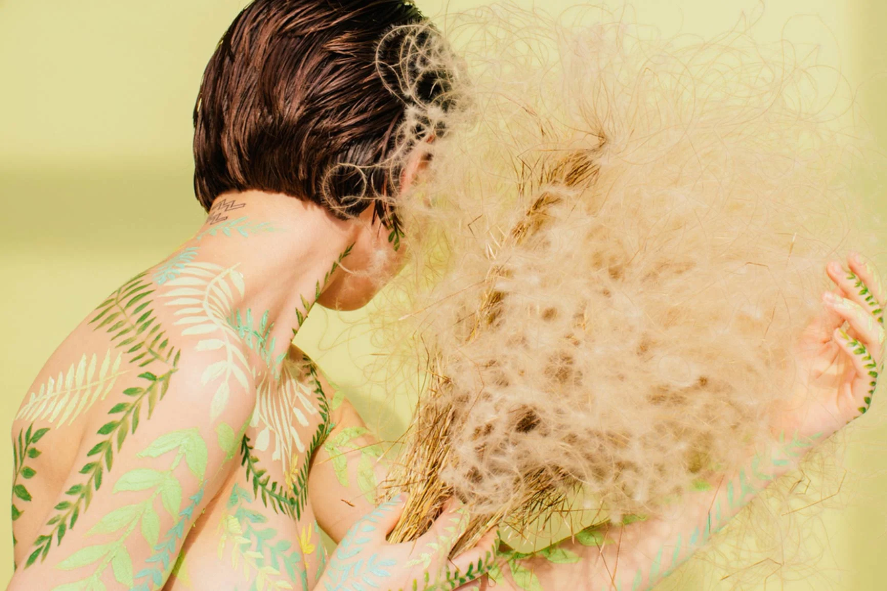 Fern-like marks on a woman's arm and back A woman, whose bare back and arm are painted with green fern leaves, holds a large, fluffy, light-colored plant umbel in front of her face.