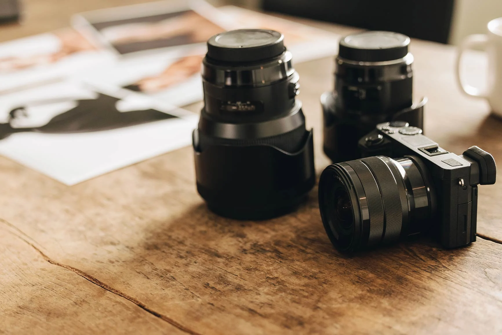 Camera and lenses on a wooden table Two camera lenses and a mirrorless camera are lying on a wooden table. In the background are out-of-focus photo prints and a white cup.
