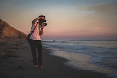 Fotograf am Strand Person fotografiert barfuß am Strand bei Sonnenuntergang, mit Kamera in den Händen.