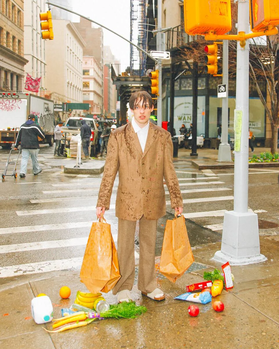 Nemo in einem braunen Anzug steht an einer nassen Straßenecke in NYC Ein ungepflegt wirkender Mann in einem schlammig-braunen Anzug steht an einer nassen Straßenecke in New York City und hält Taschen, während Lebensmittel um ihn herum auf dem Boden verstreut liegen.