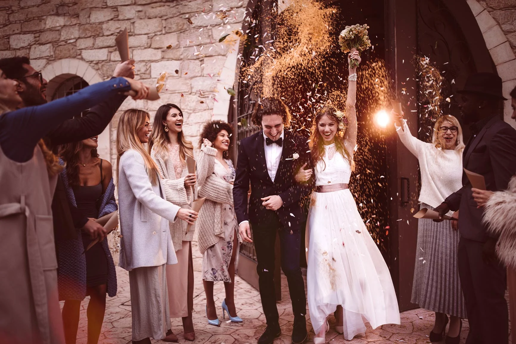 The bride and groom surrounded by guests The bride and groom stand in front of a stone archway, surrounded by guests throwing confetti.