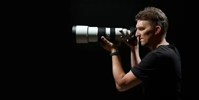 Les types d'objectifs et leur utilisation 5 Une personne portant un t-shirt noir tient un appareil photo avec un grand téléobjectif, sur un fond sombre.