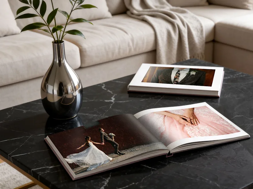 SEA / SEO LP | Wedding Photo Book | Product Selector Section (US, BE, IT, ES, EU) | Media #1 Dos álbumes de fotos de boda abiertos sobre una mesa de centro de mármol negro, junto a un jarrón plateado, con un sofá color crema al fondo.