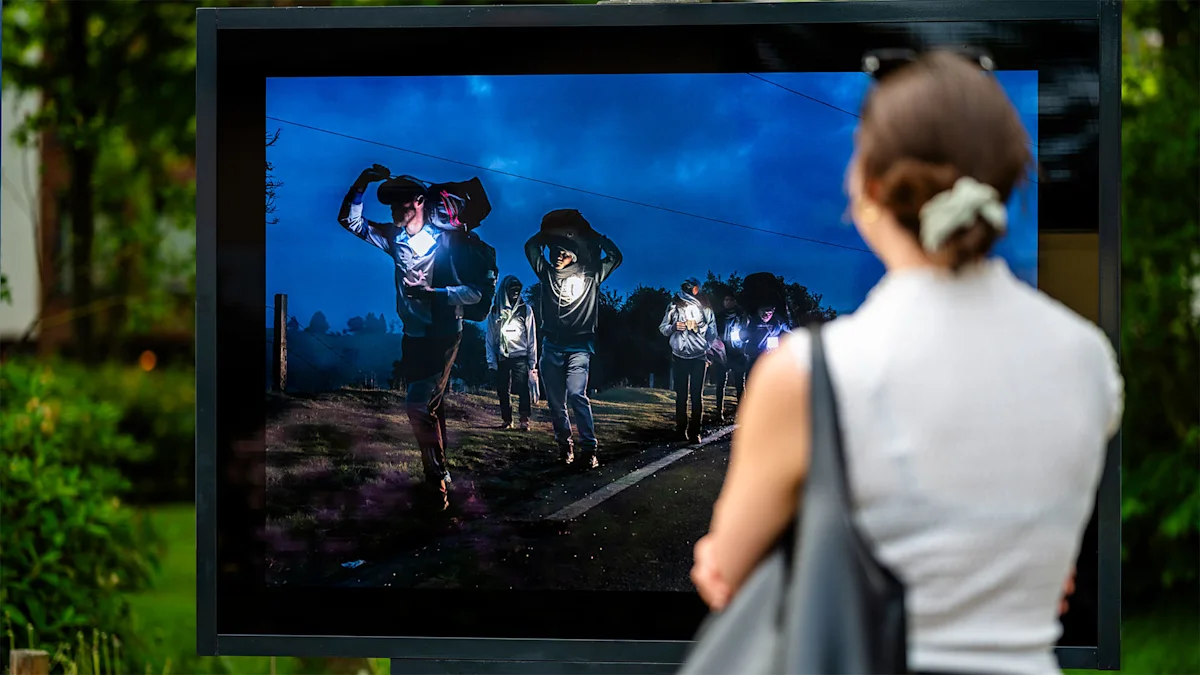 Rubén Salgado Outdoor Exhibition in Zingst A woman views an outdoor photo exhibit showing people walking at night with lights, set against a dark blue sky.