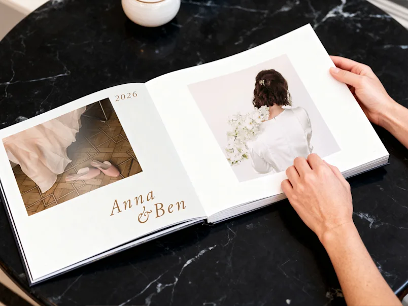 SEA / SEO LP | Wedding Photo Book | Product Selector Section (US, BE, IT, ES, EU) | Media #2 Hands holding open a wedding photo book labeled Anna & Ben 2026, showing bridal shoes and a bride with white flowers.