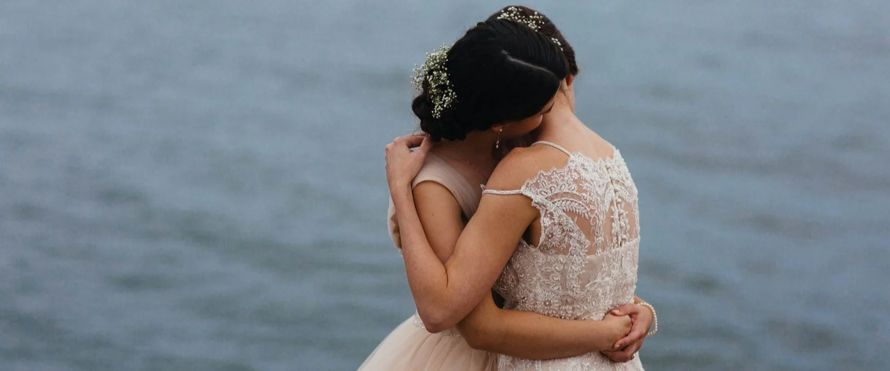 Two women in elegant lace dresses are hugging each other in front of a body of water.