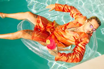 Bill Kaulitz in orangefarbener Kleidung, der im Pool entspannt Mann in orangefarbenem Outfit und mit pinken Sonnenbrillen entspannt auf einer Pool-Luftmatratze und hält einen pinken Cocktail mit einer Zitronenscheibe.